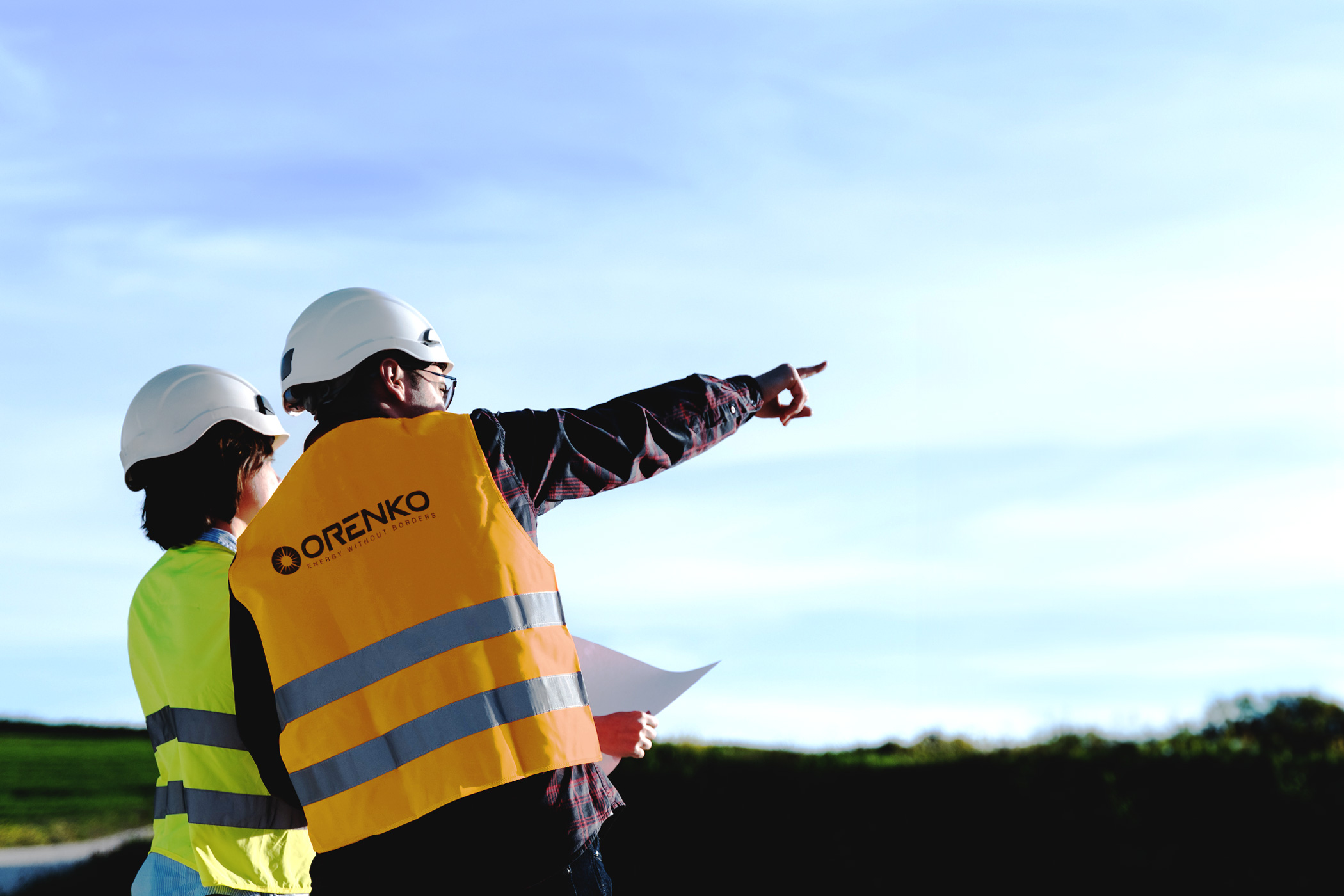 Two construction workers in helmets and safety vests looking and pointing outdoors with papers in hand.