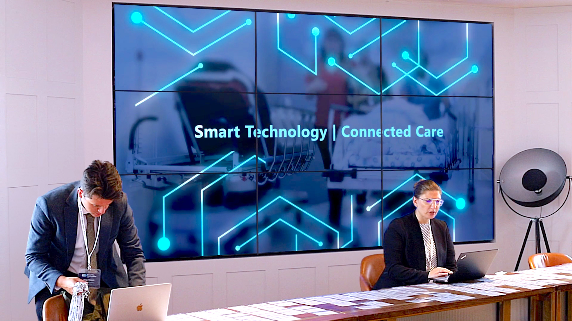 Man and woman working on laptops at table with cards; large screen reads Smart Technology | Connected Care.