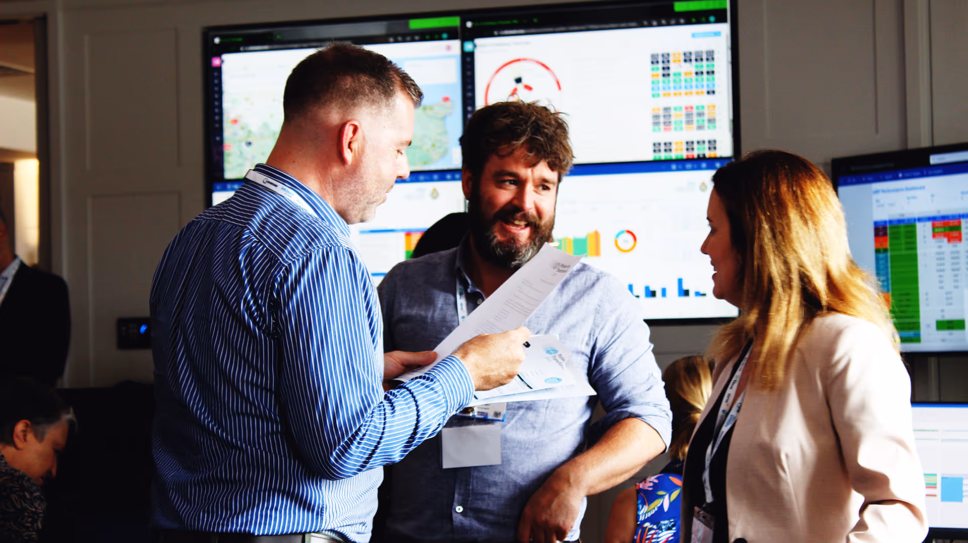 Three professionals discussing documents in front of monitors displaying data charts and maps.