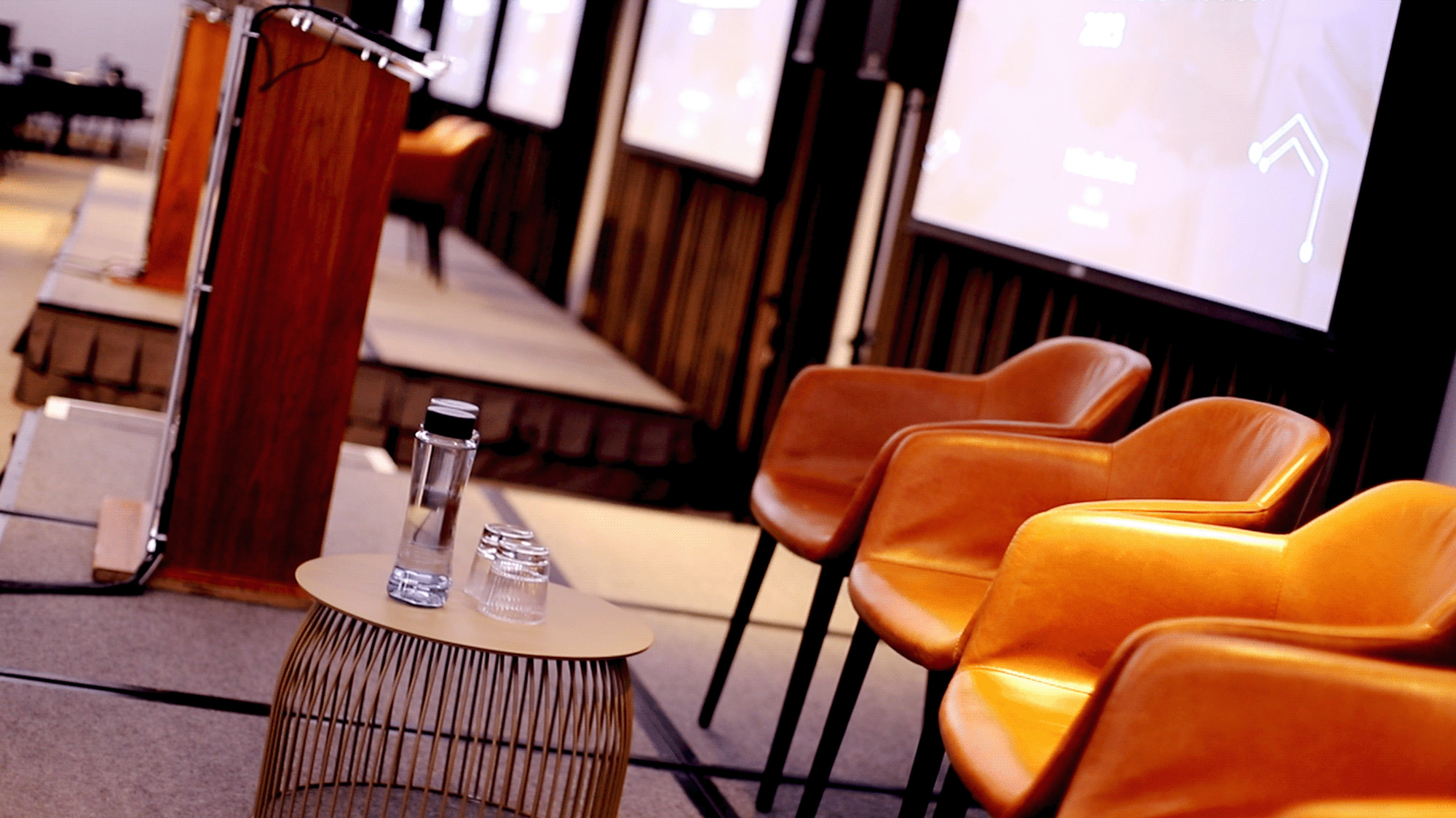 Stage with wooden lectern, three empty orange chairs, and a small round table with water bottle and glasses.