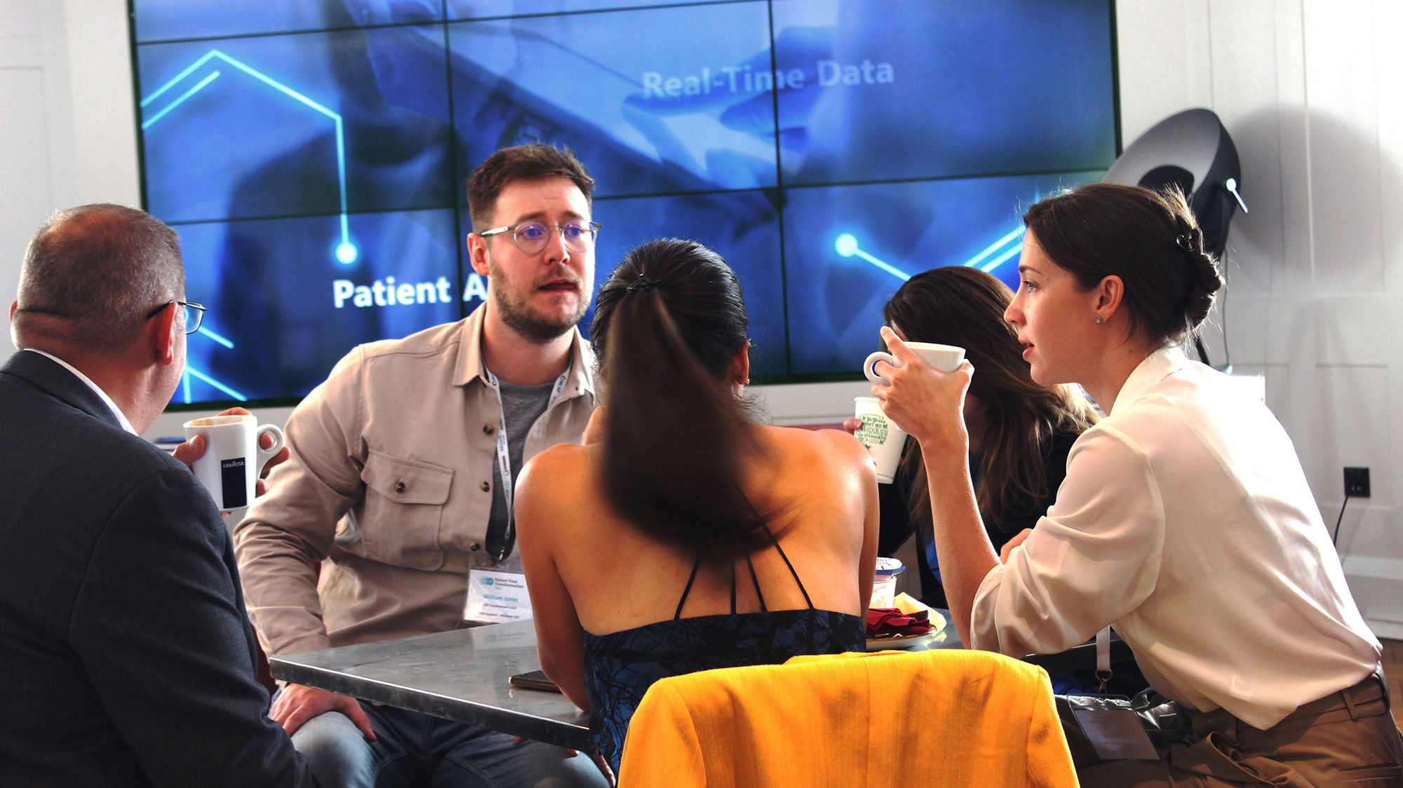 Four people sitting around a table having a discussion with coffee cups in front of a screen showing data.