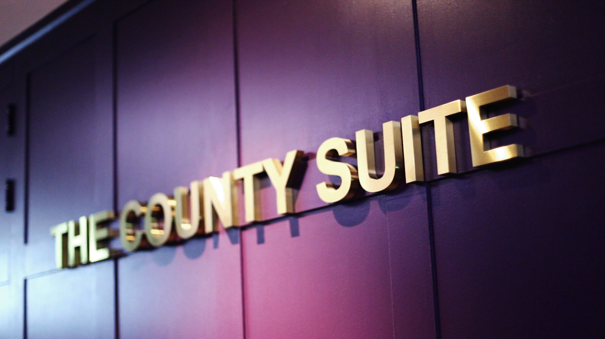 Gold letters spelling 'THE COUNTY SUITE' on a purple paneled wall with shadows.