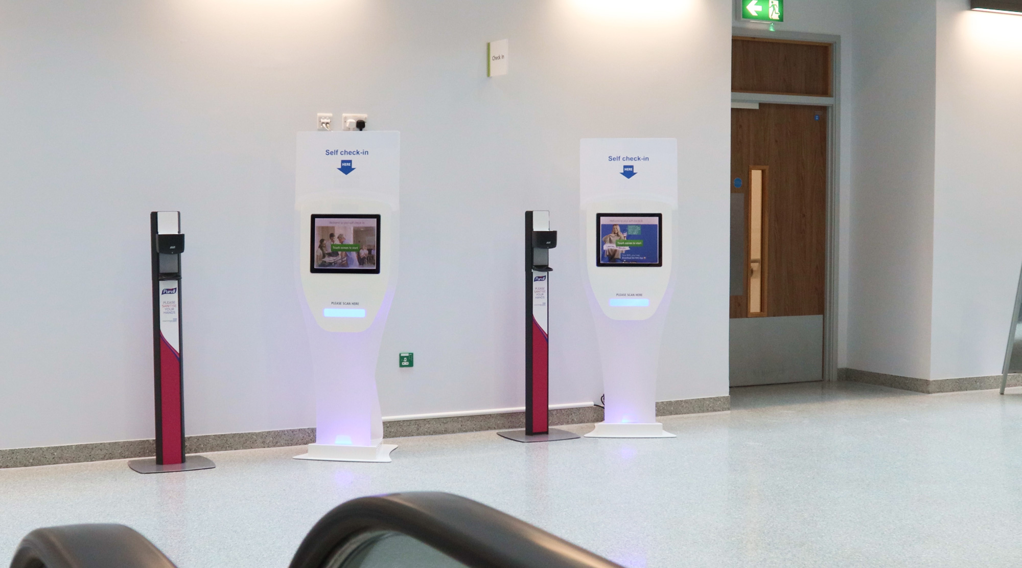 Two self check-in kiosks with hand sanitizer stations in a spacious, well-lit lobby area.