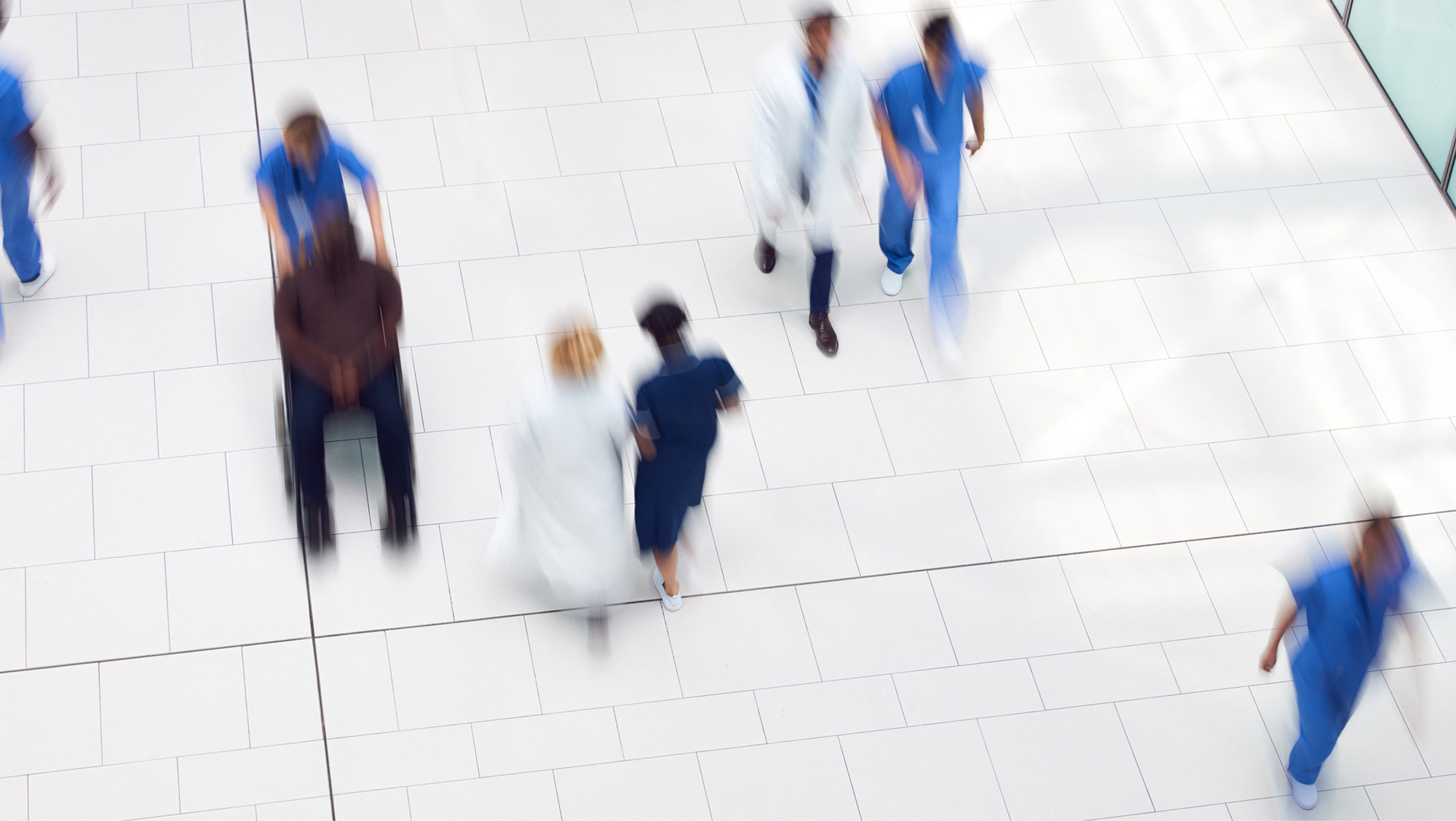 Blurred overhead view of diverse healthcare workers and a patient in a wheelchair in a hospital corridor.