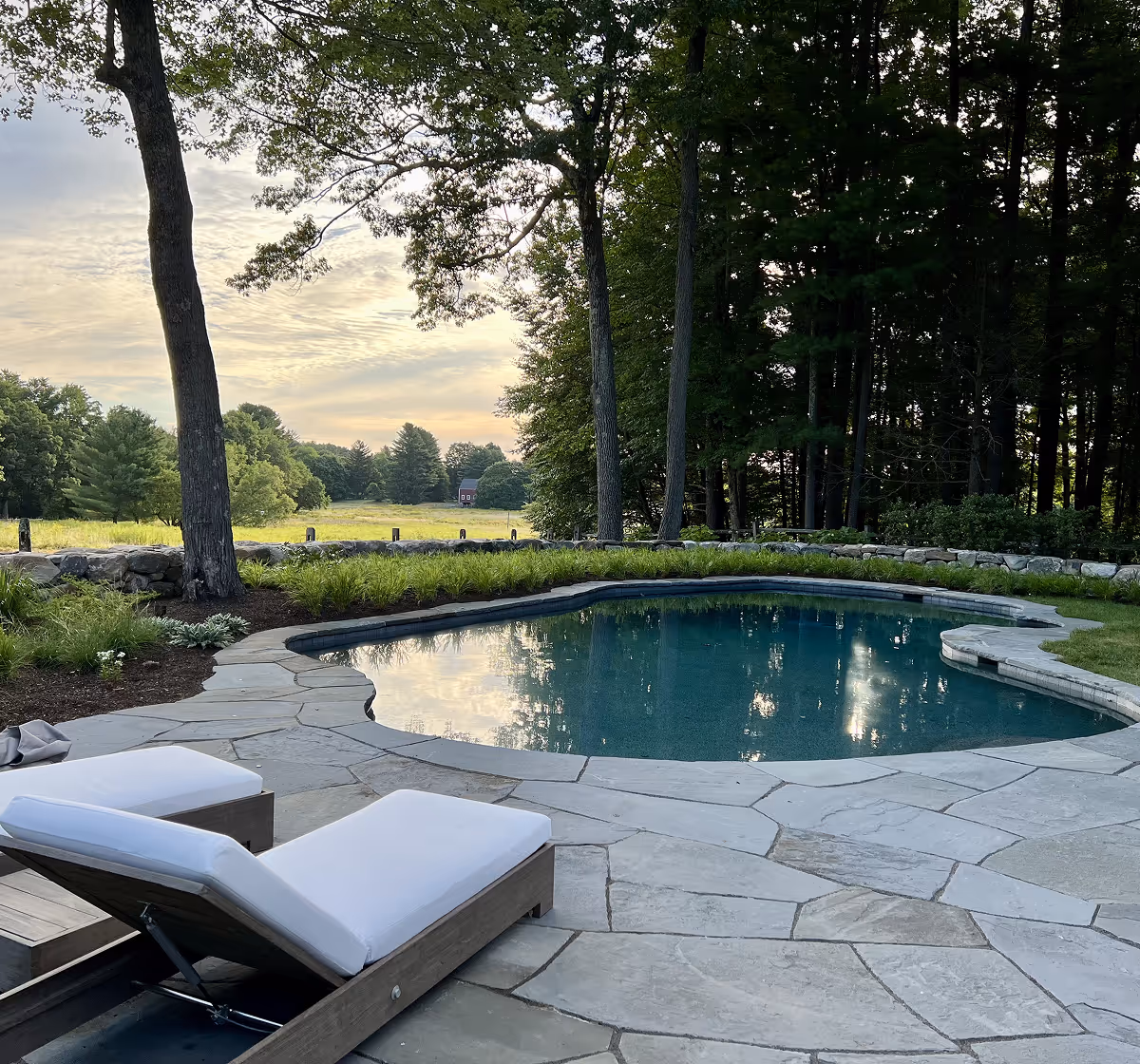 Outdoor swimming pool surrounded by stone deck and greenery with two white-cushioned lounge chairs in the foreground under trees at sunset.