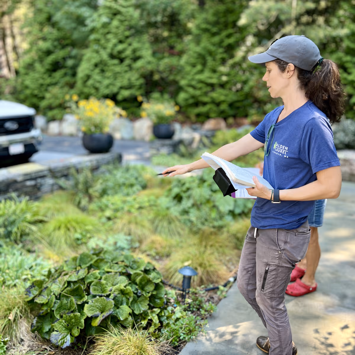Woman in a blue Walden Laurel t-shirt and gray cap pointing at plants while holding notes in a garden.