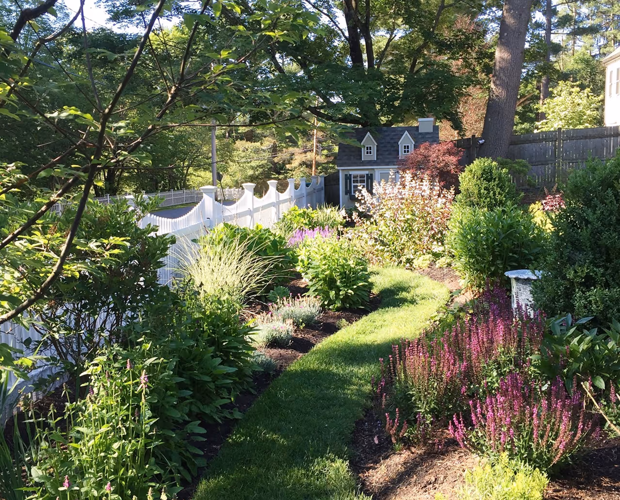Garden with green grass path, colorful flowering plants, shrubs, a white fence, and a small playhouse in the background.
