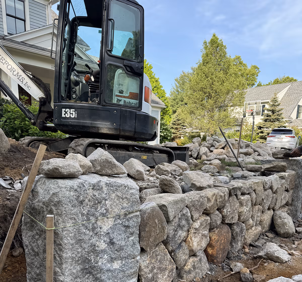 Construction site with a Bobcat excavator and a partially built stone retaining wall in a residential area.