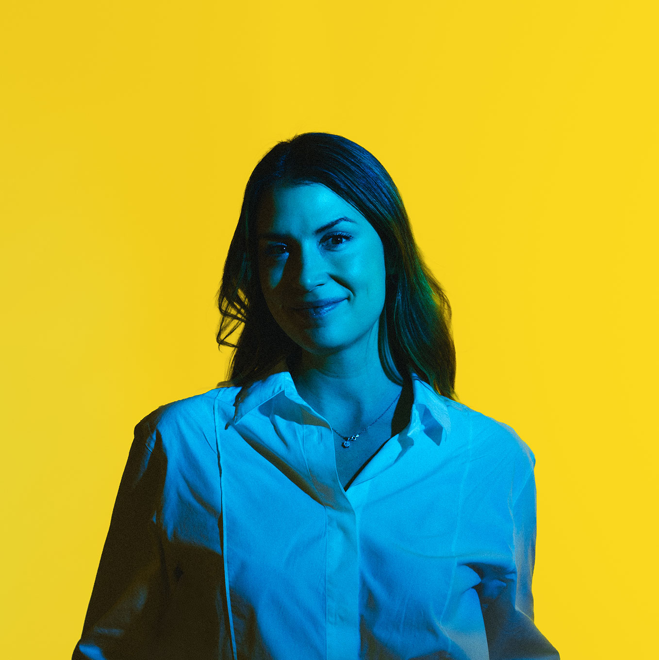 Young woman with long hair wearing a white shirt, lit by blue light, against a solid yellow background.