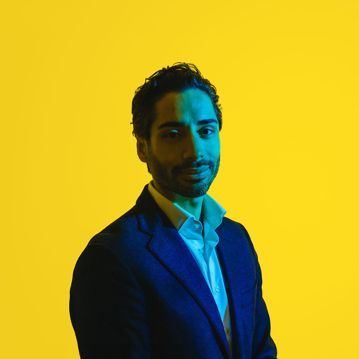 Man in a dark suit jacket and white shirt with blue and yellow lighting on a yellow background.