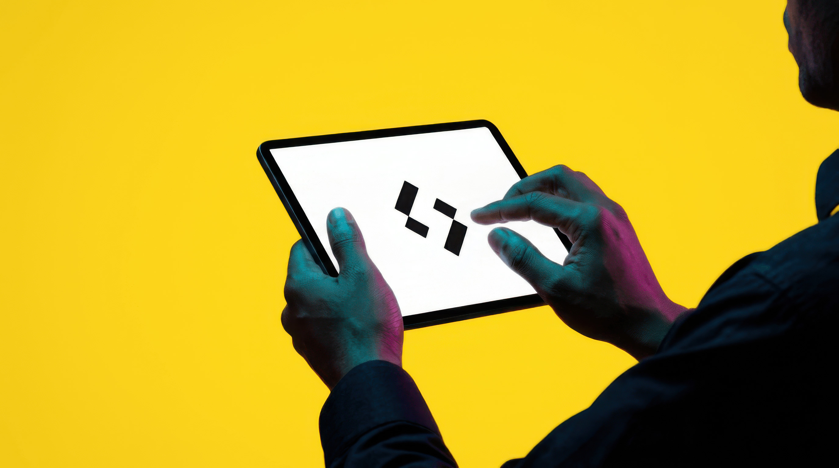 Person holding and interacting with a tablet displaying a black geometric logo on a white screen against a yellow background.