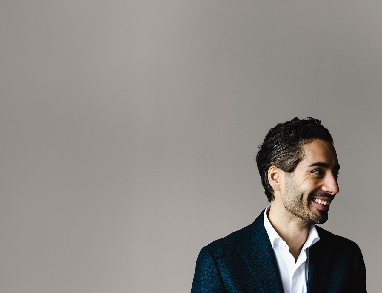 Smiling man with dark hair wearing a white shirt and dark blazer looking to the right against a plain gray background.