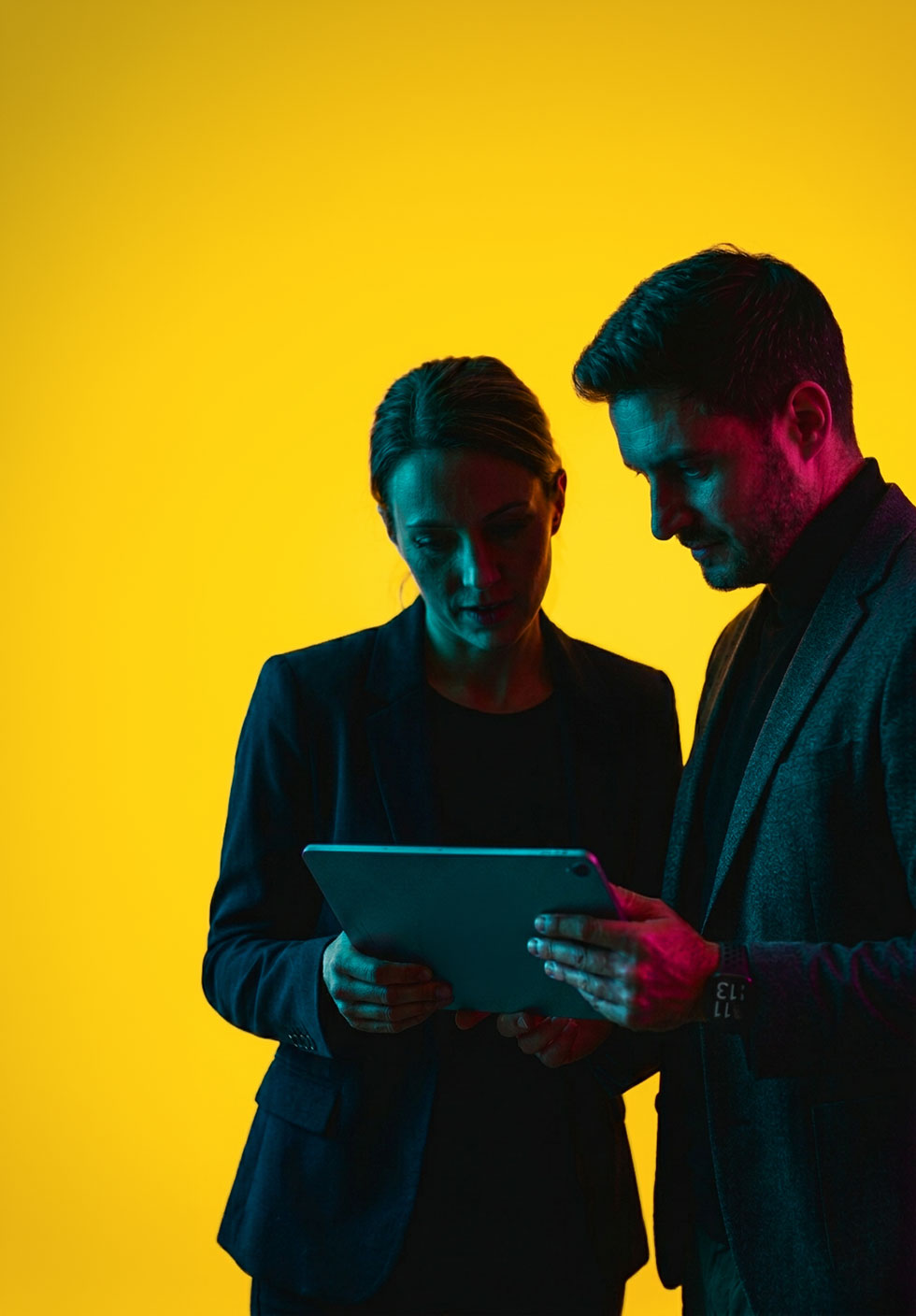 Man and woman in business attire looking at a tablet against a yellow background.