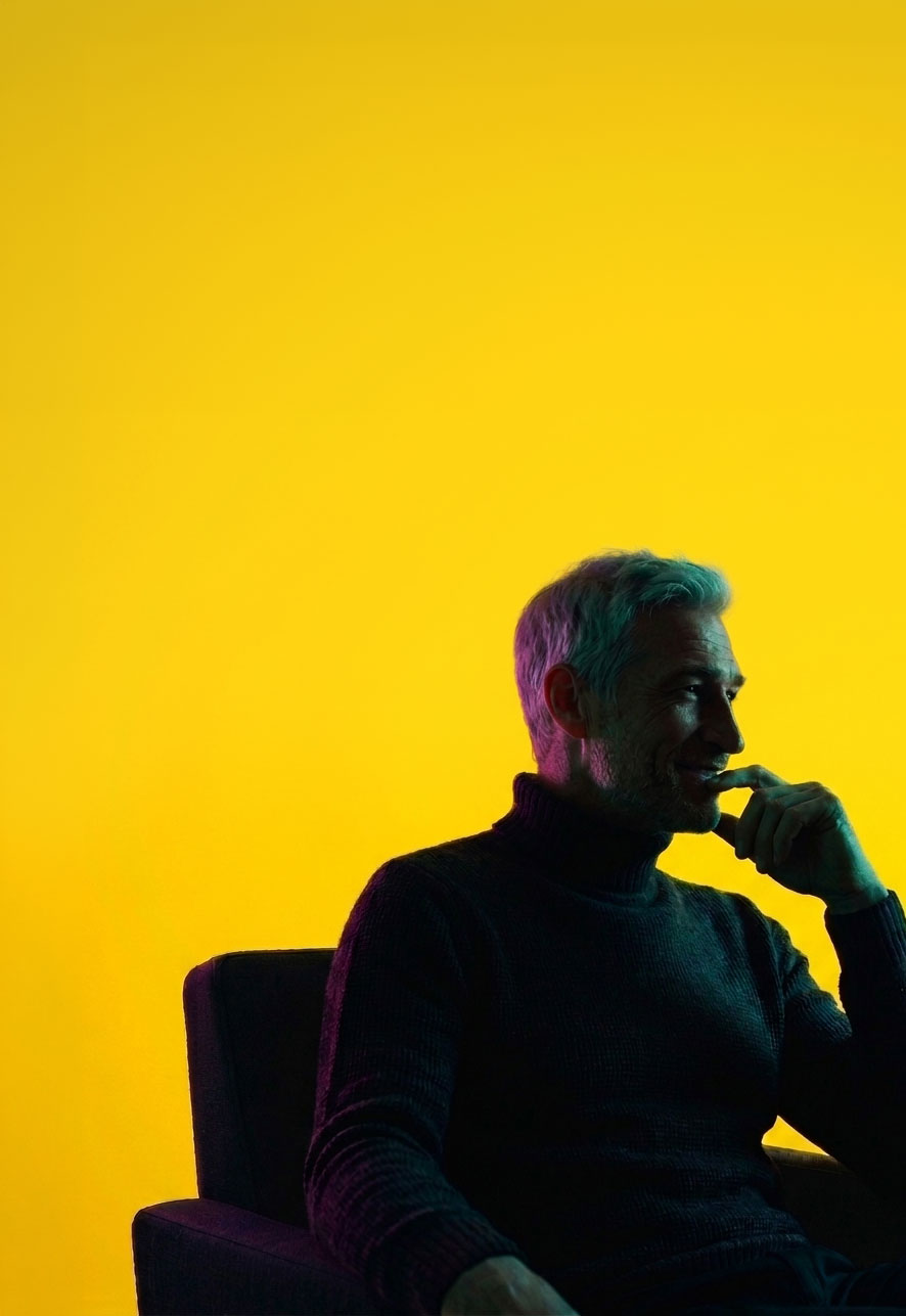 Silhouette of a man with gray hair and beard sitting on a chair against a bright yellow background, resting his chin on his hand thoughtfully.