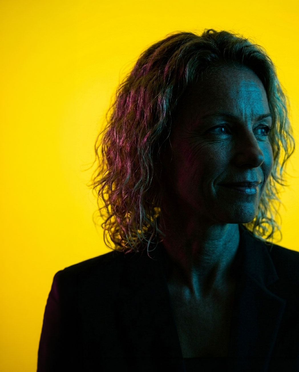Profile of a woman with curly hair illuminated by blue and purple lighting against a bright yellow background.