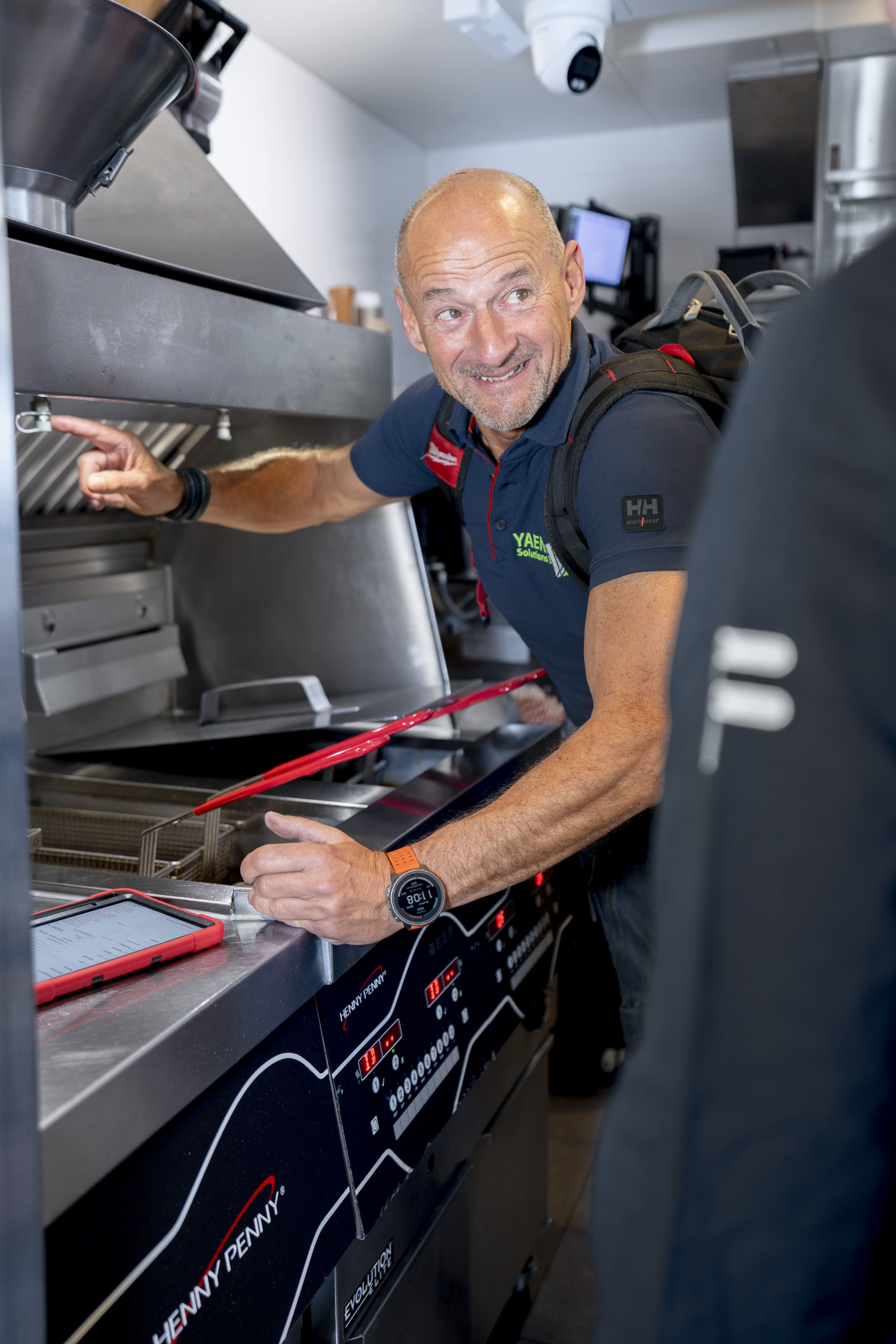 Man met rugzak wijst naar keukenapparatuur in professionele keukenomgeving.