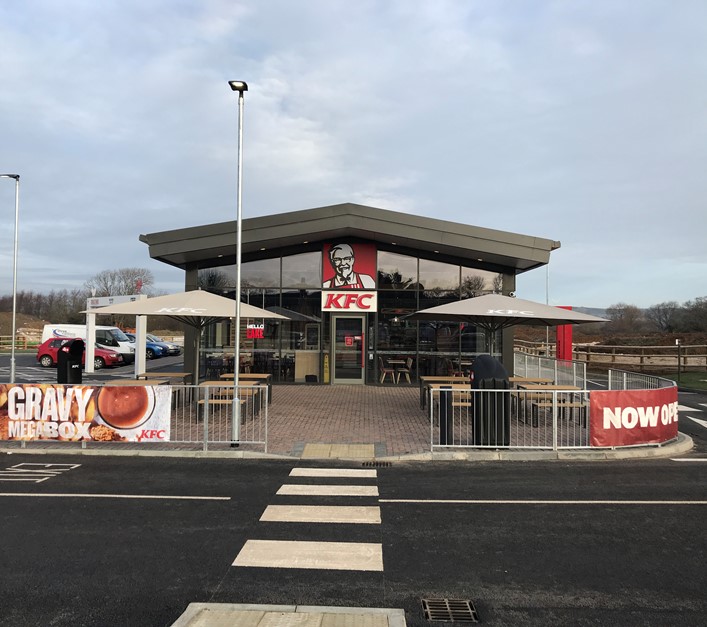 Vooraanzicht van een KFC-restaurant met buitenterrassen, parasols en een oversteekplaats naar de ingang.