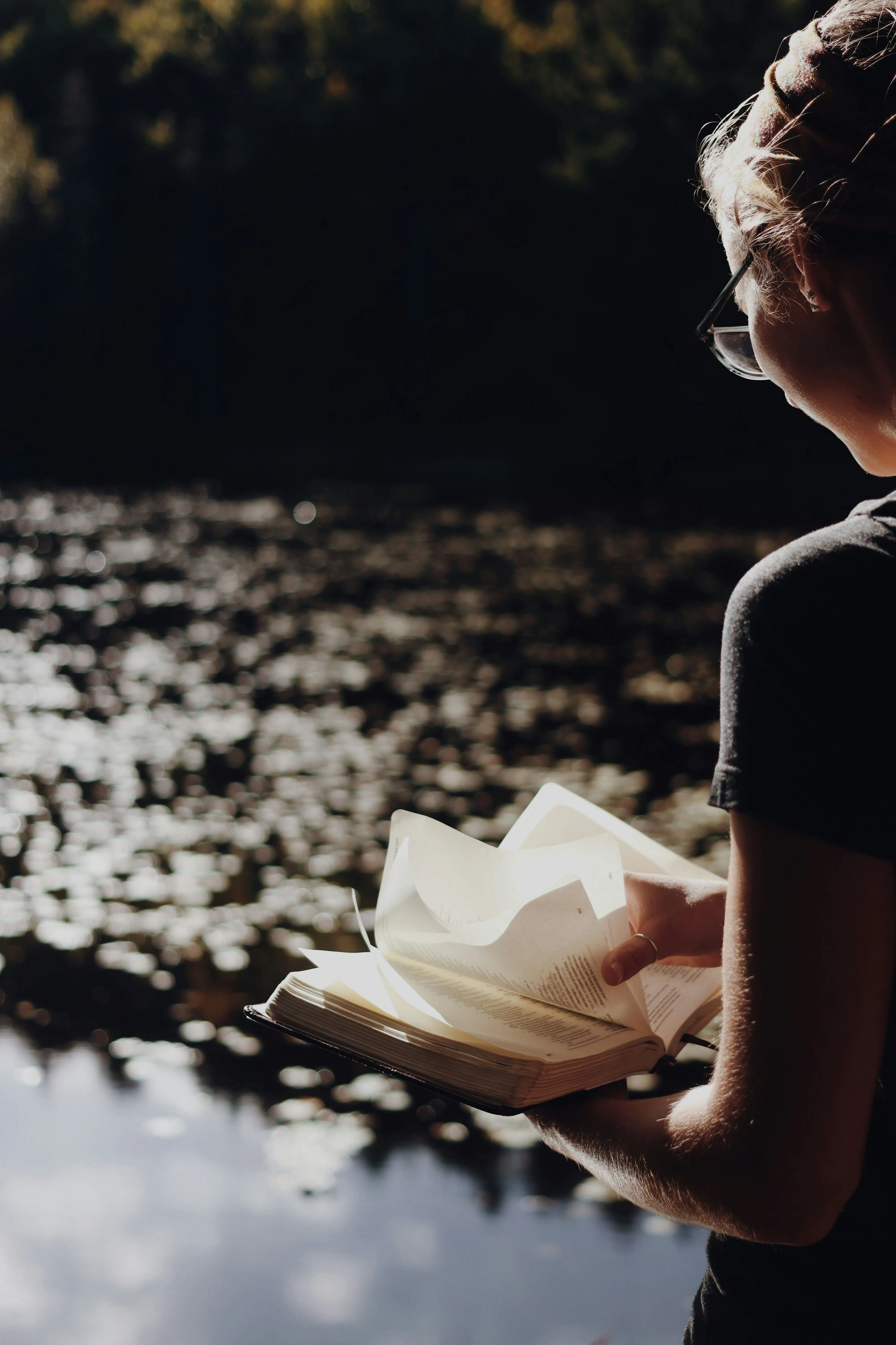 Une personne avec lunettes lisant un livre près d’une surface d’eau miroitante, arrière‑plan flou.