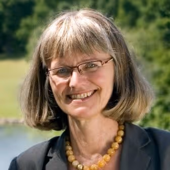 Smiling woman with shoulder-length gray hair and glasses, wearing a dark blazer and a yellow beaded necklace, outdoors with greenery in the background.