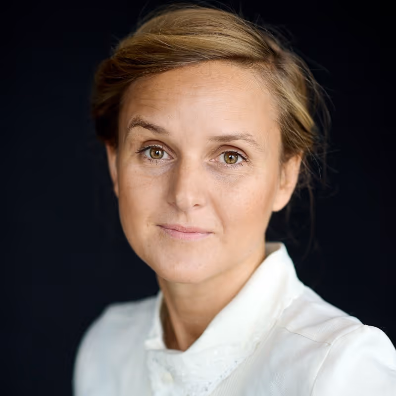 Portrait of a woman with light brown hair and hazel eyes wearing a white top against a black background.
