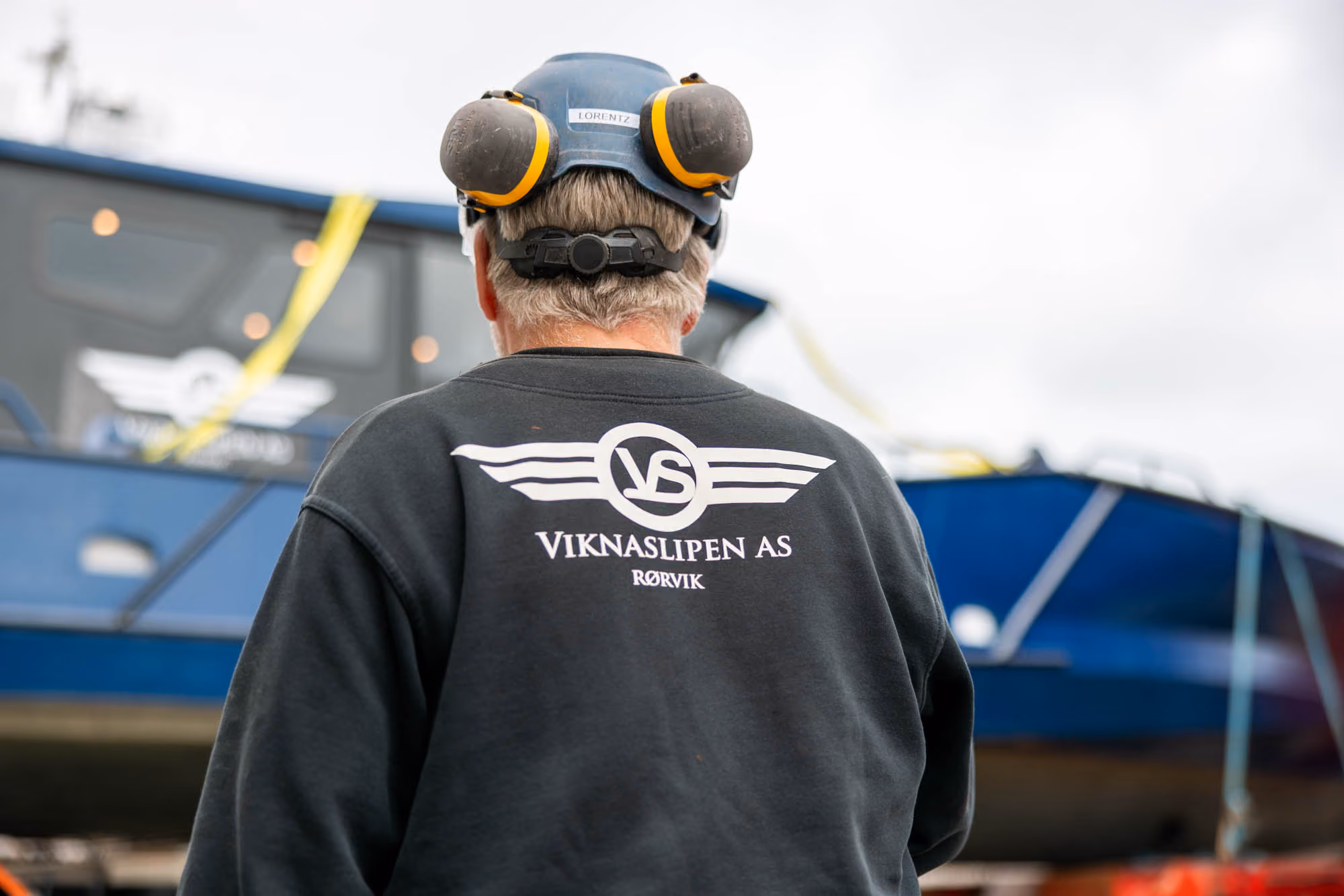 Mann med hjelm og hørselsvern står foran en blå båt med logoen Viknaslipen AS Rørvik på genseren.
