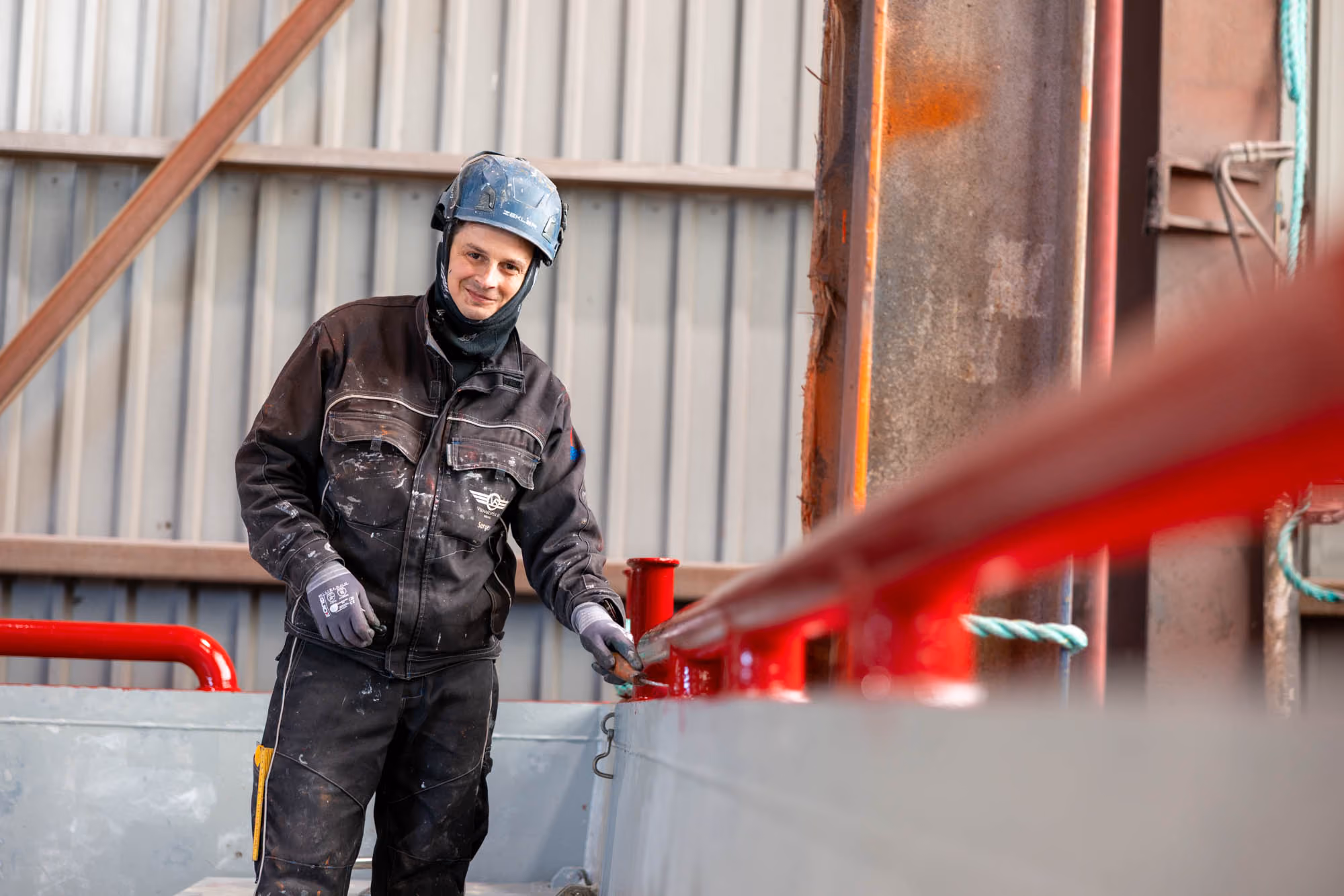Bygningsarbeider i svart arbeidstøy og hjelm holder en rød metallrekkverk inne i en industribygg.