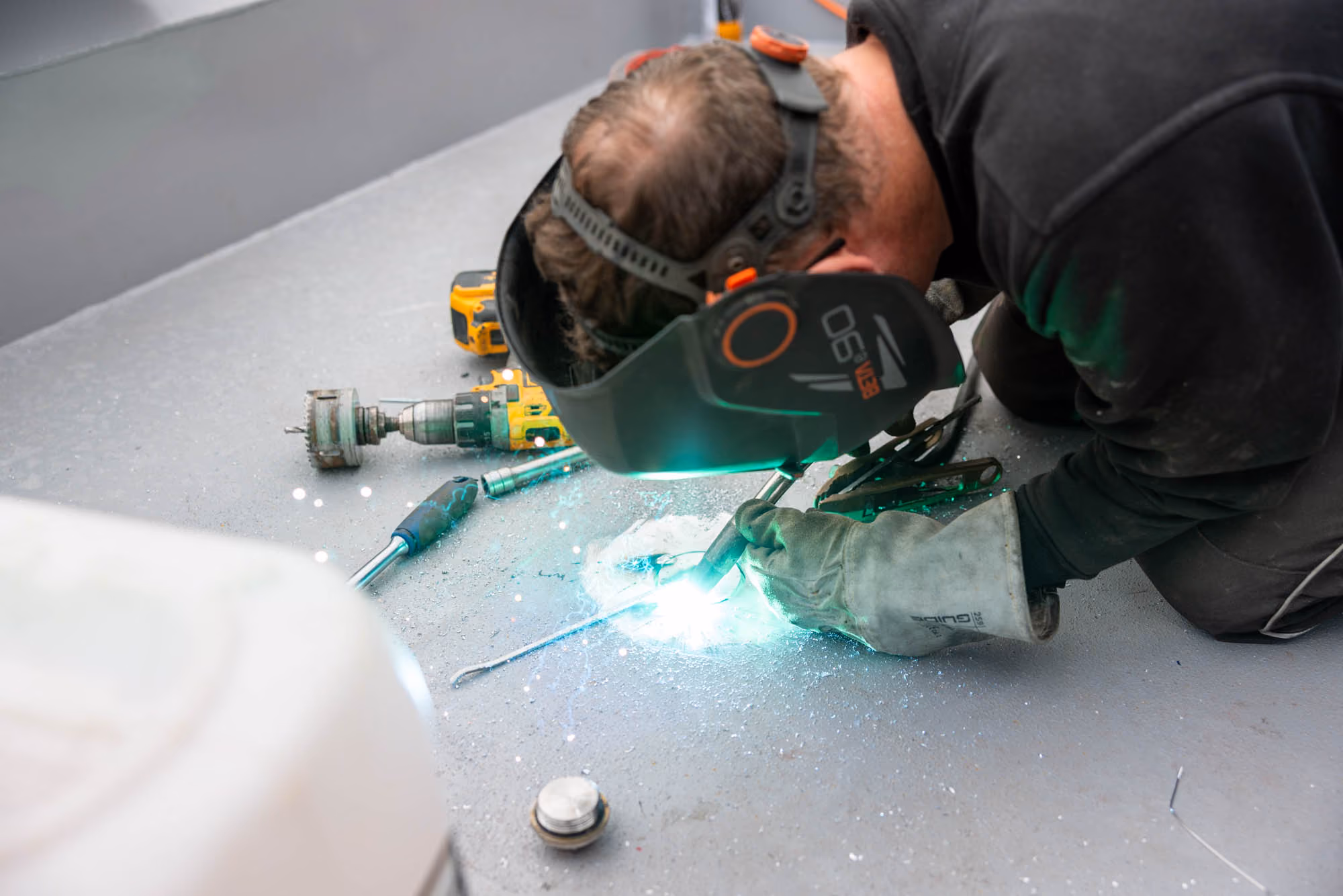 Person welding metal on a floor, wearing protective gloves and a welding helmet.