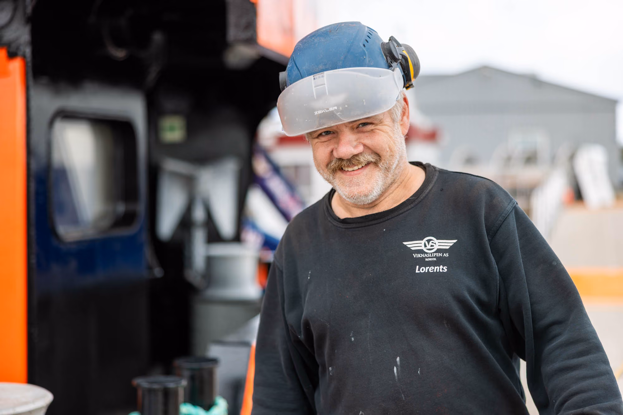 Smilende byggherre med blå hjelm og vernebriller foran industrianlegg.
