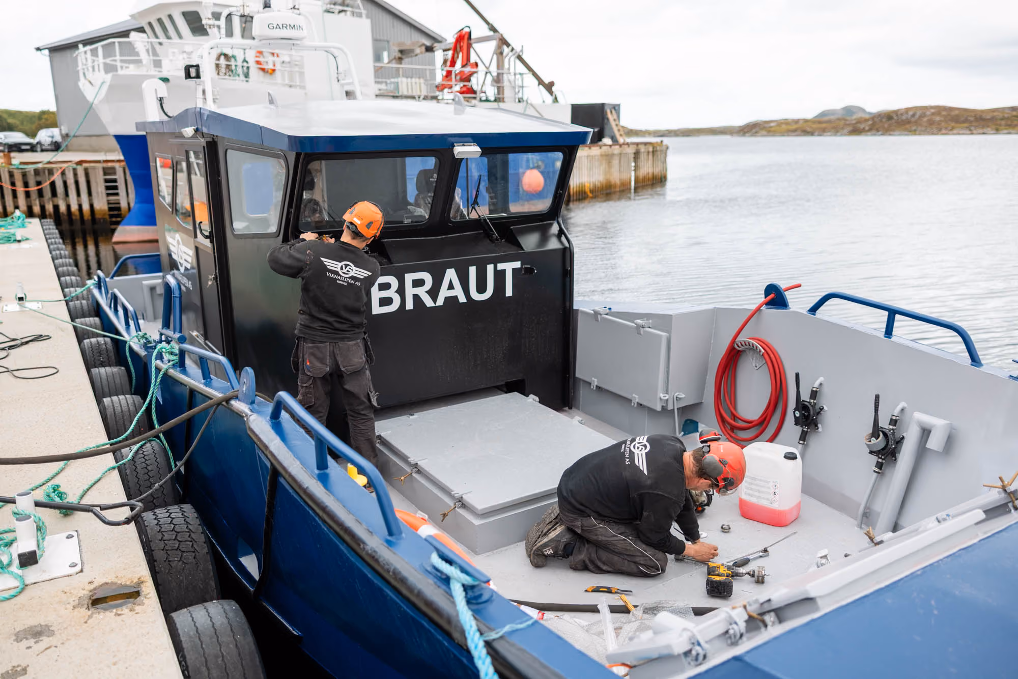 To mennelige arbeidere fester utstyr på en svart og blå båt ved en kai.