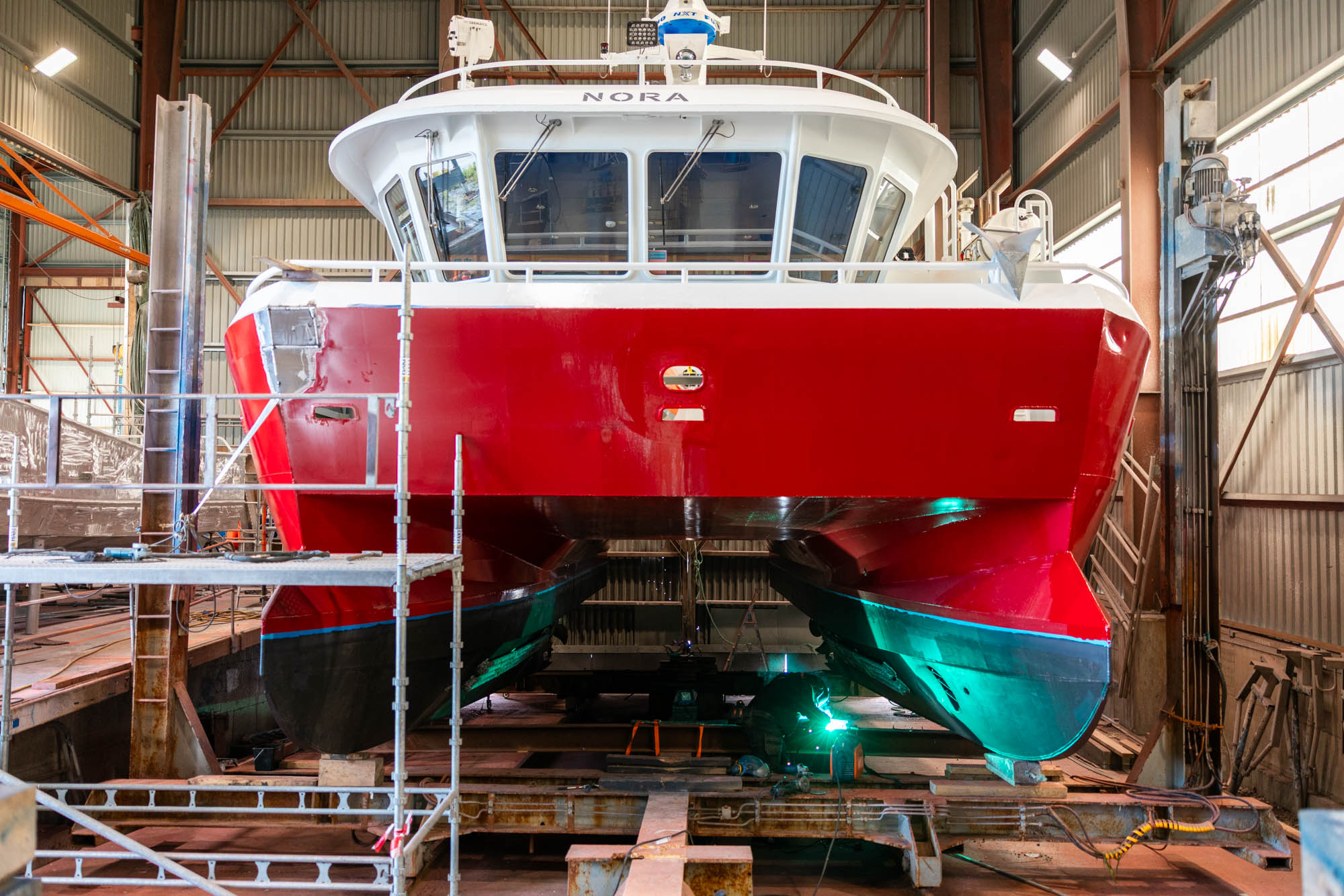 Rød og hvit katamaran i verft med arbeidsperson som sveiser under båten.