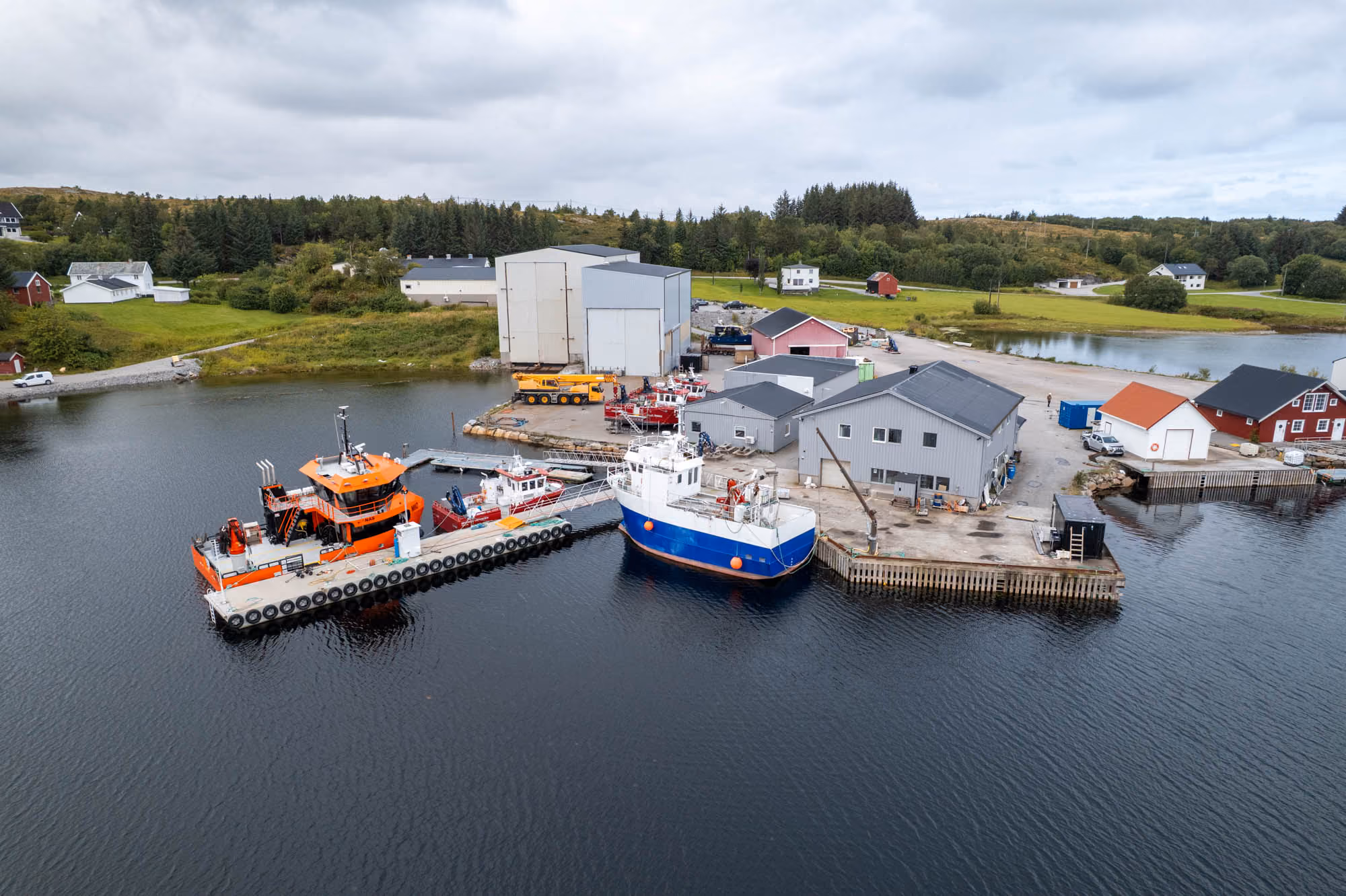 Liten havn med brygge, flere båter og industribygg ved kysten omkranset av gress og trær.
