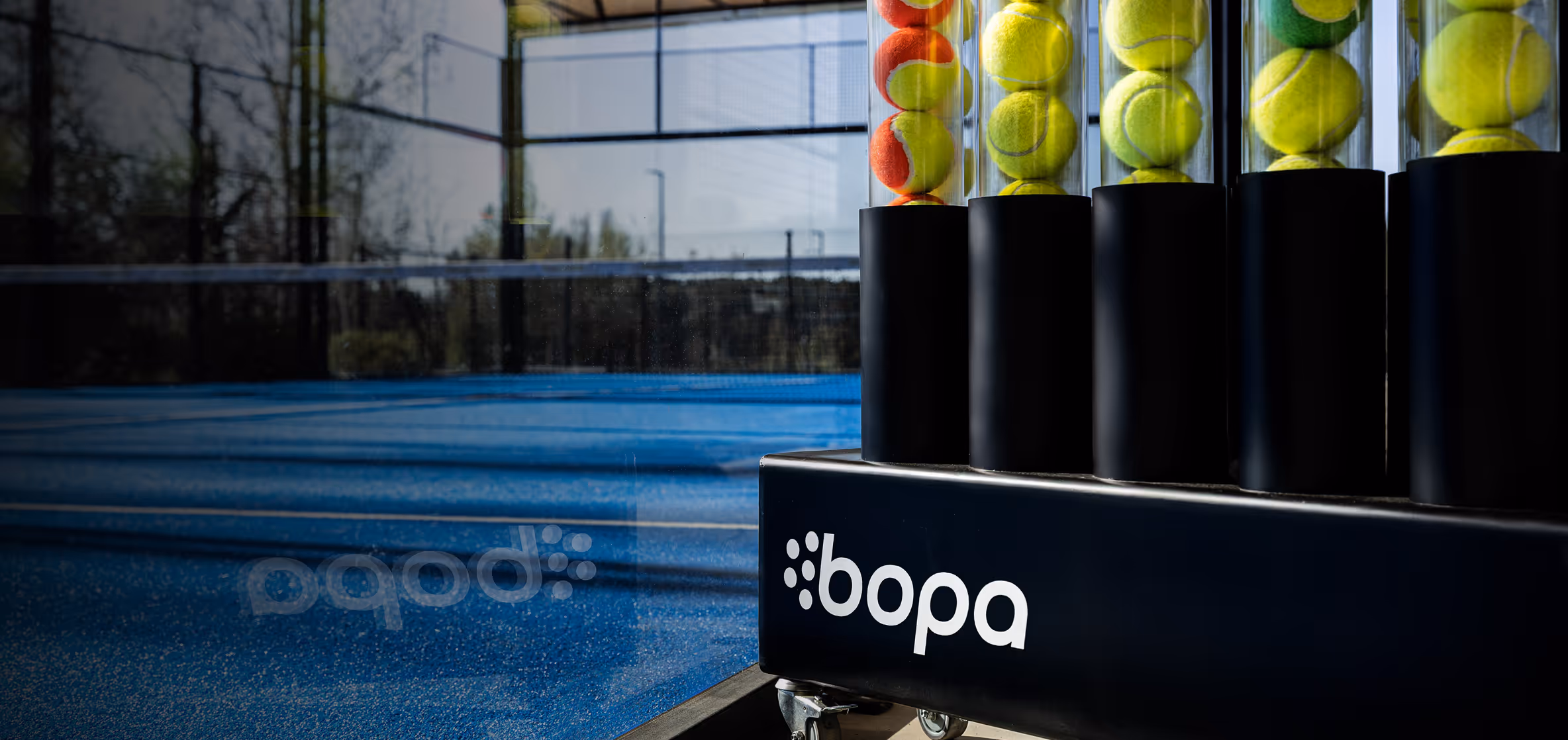 Blue padel tennis court with a ball holder containing yellow and orange tennis balls marked with 'bopa'.