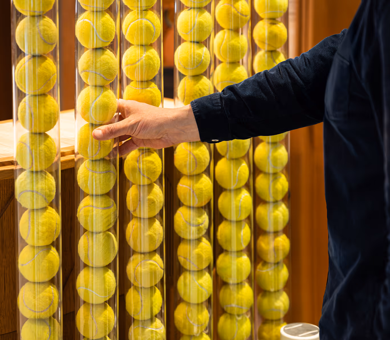Person's hand retrieving a tennis ball from a vertical clear tube filled with yellow tennis balls.