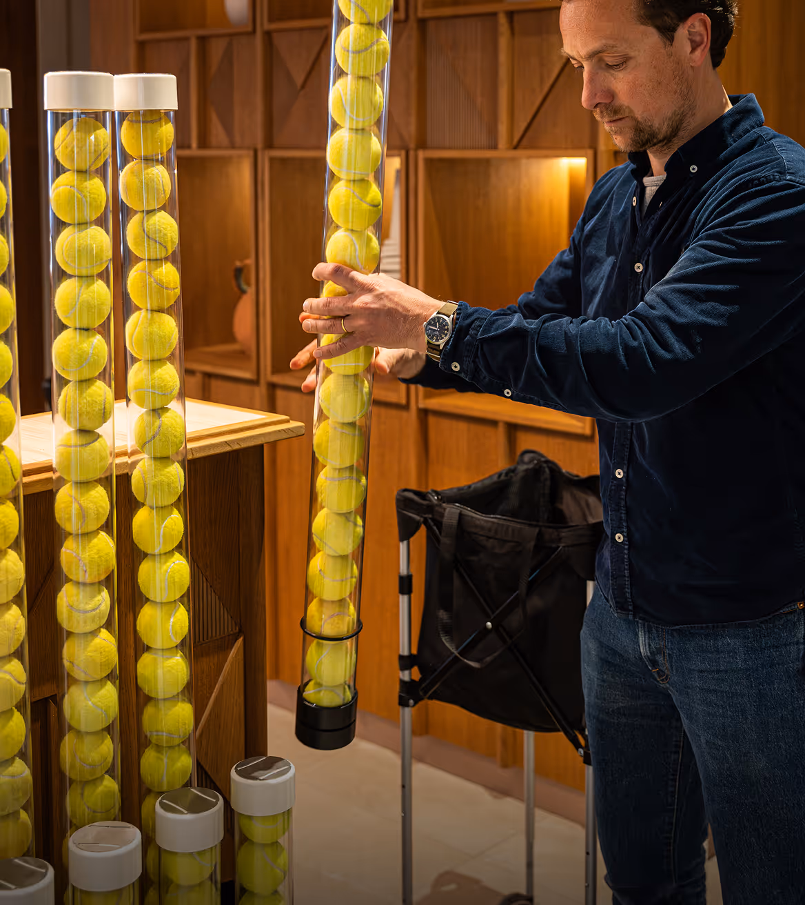 Man arranging yellow tennis balls inside a transparent tube in a wooden-paneled room.