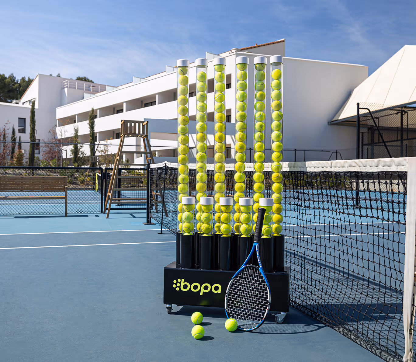 Tennis ball dispenser filled with yellow tennis balls and a blue tennis racket leaning on it near the tennis net on an outdoor court.