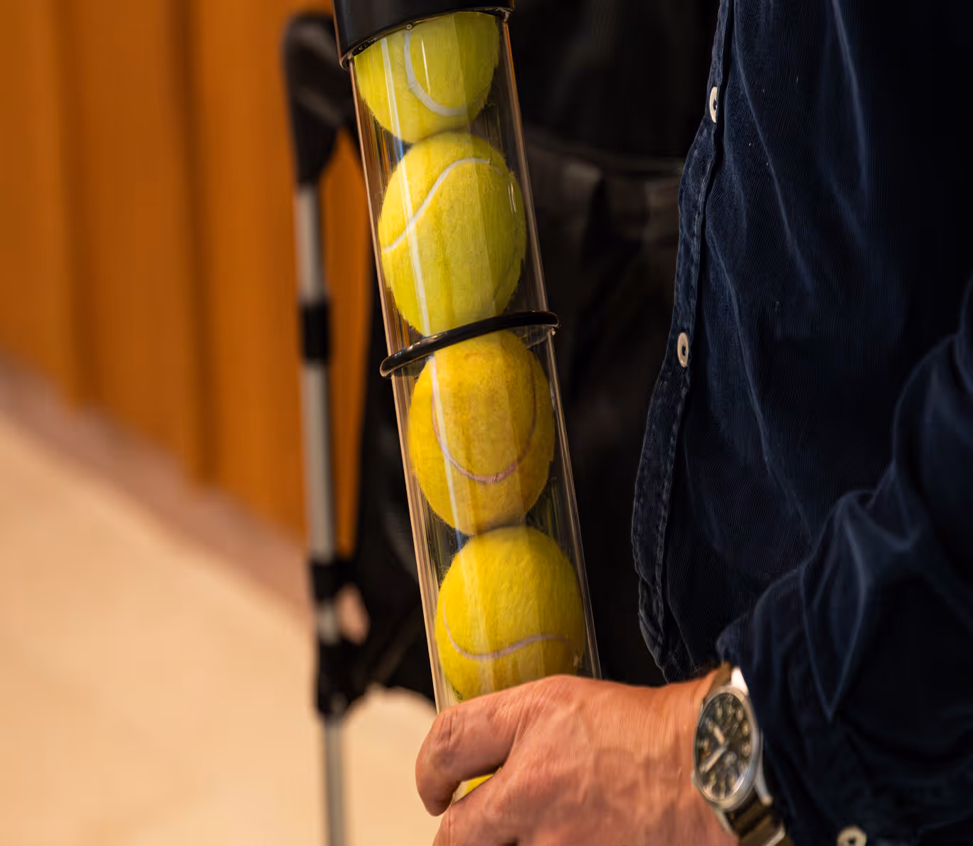 Close-up of a person holding a transparent tube containing four yellow tennis balls with a golf bag and club in the background.