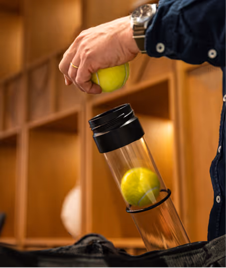 Hand holding a tennis ball above a transparent tennis ball container partially inside a bag, with wooden shelves in the background.