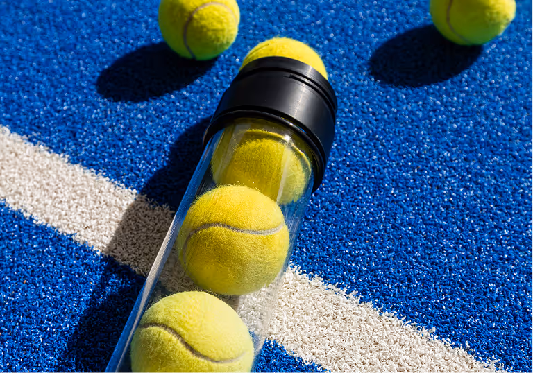 Several yellow tennis balls on a blue tennis court, with some inside a clear plastic container with a black lid.