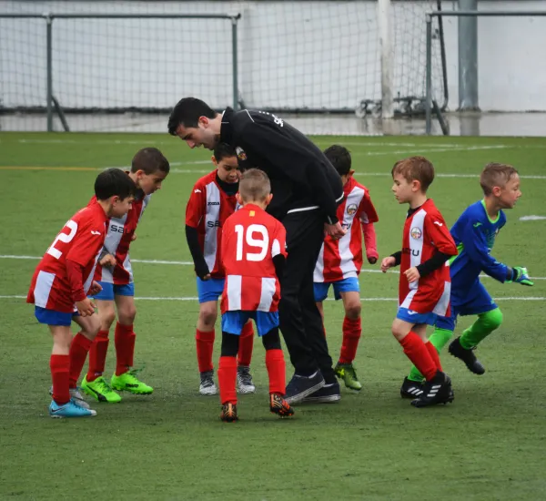 Young children dressed mostly in red and white football kits gather round a coach on an astroturf pitch