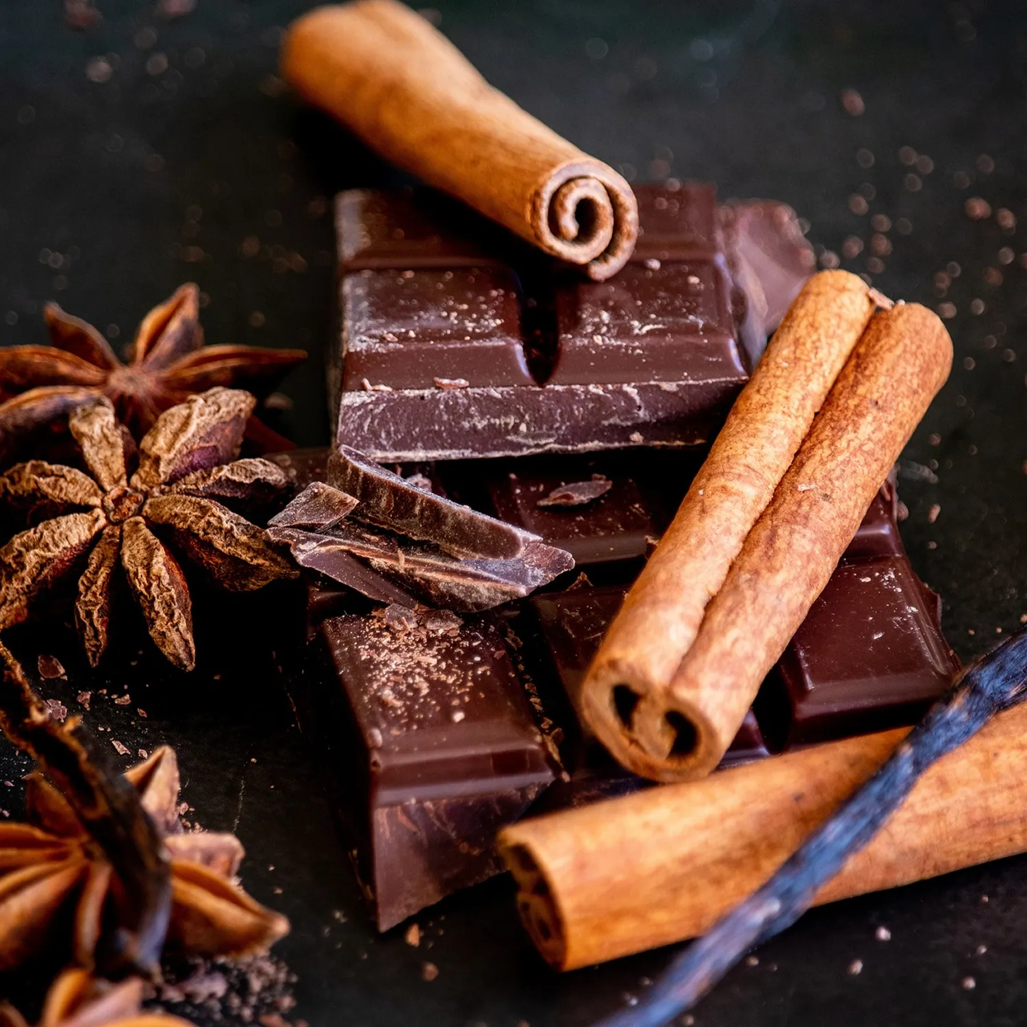 Dark chocolate pieces surrounded by cinnamon sticks and star anise on a dark surface.