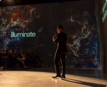 A man in a dark suit holding a microphone stands on a stage illuminated by warm lights, speaking to an audience seated on the left. Behind him is a large screen displaying a colorful nebula-like pattern with the word 'Illuminate' projected on it.