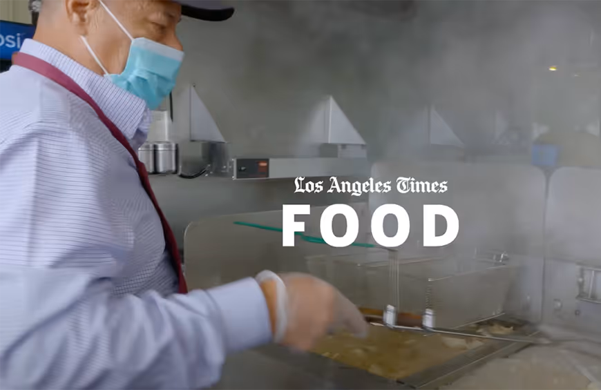Person wearing a mask and cap cooking food in a deep fryer with steam rising, with 'Los Angeles Times FOOD' text overlay.