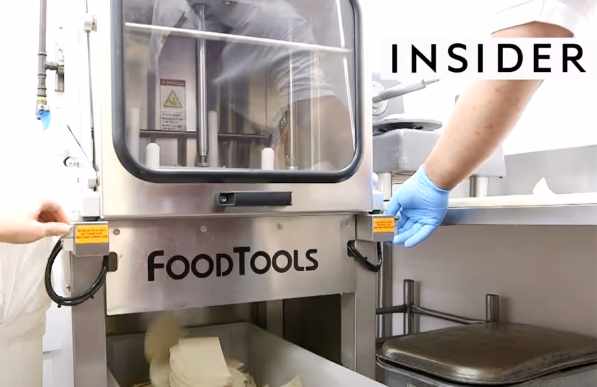 Two hands, one wearing a blue glove, operating a stainless steel FOODTOOLS machine in a kitchen with slices of soft cheese or tofu dropping into a container below.