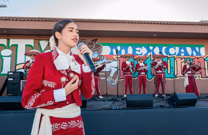 Female mariachi singer in traditional red outfit performing with a mariachi band on an outdoor stage in front of a colorful mural.