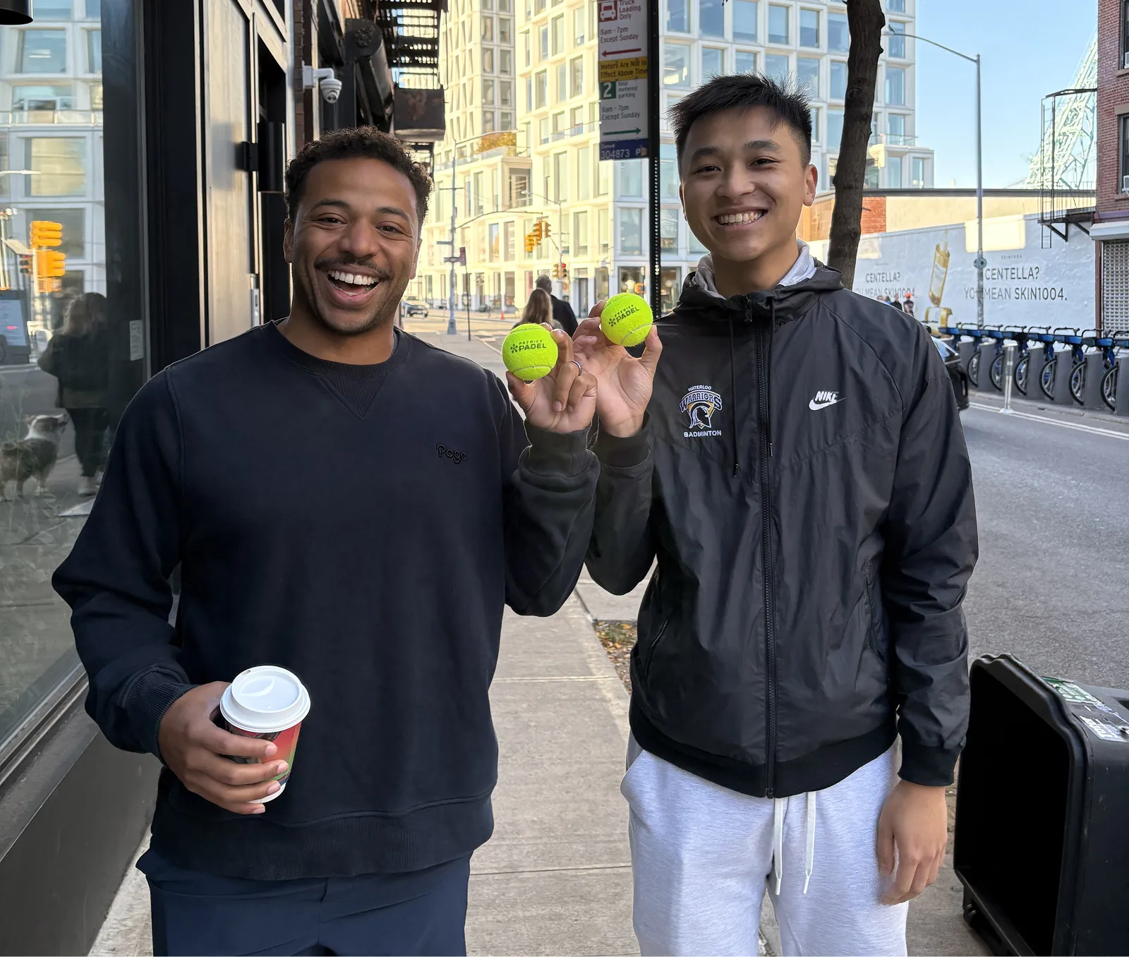 Two men smiling on a city sidewalk, each holding a bright yellow padel ball.