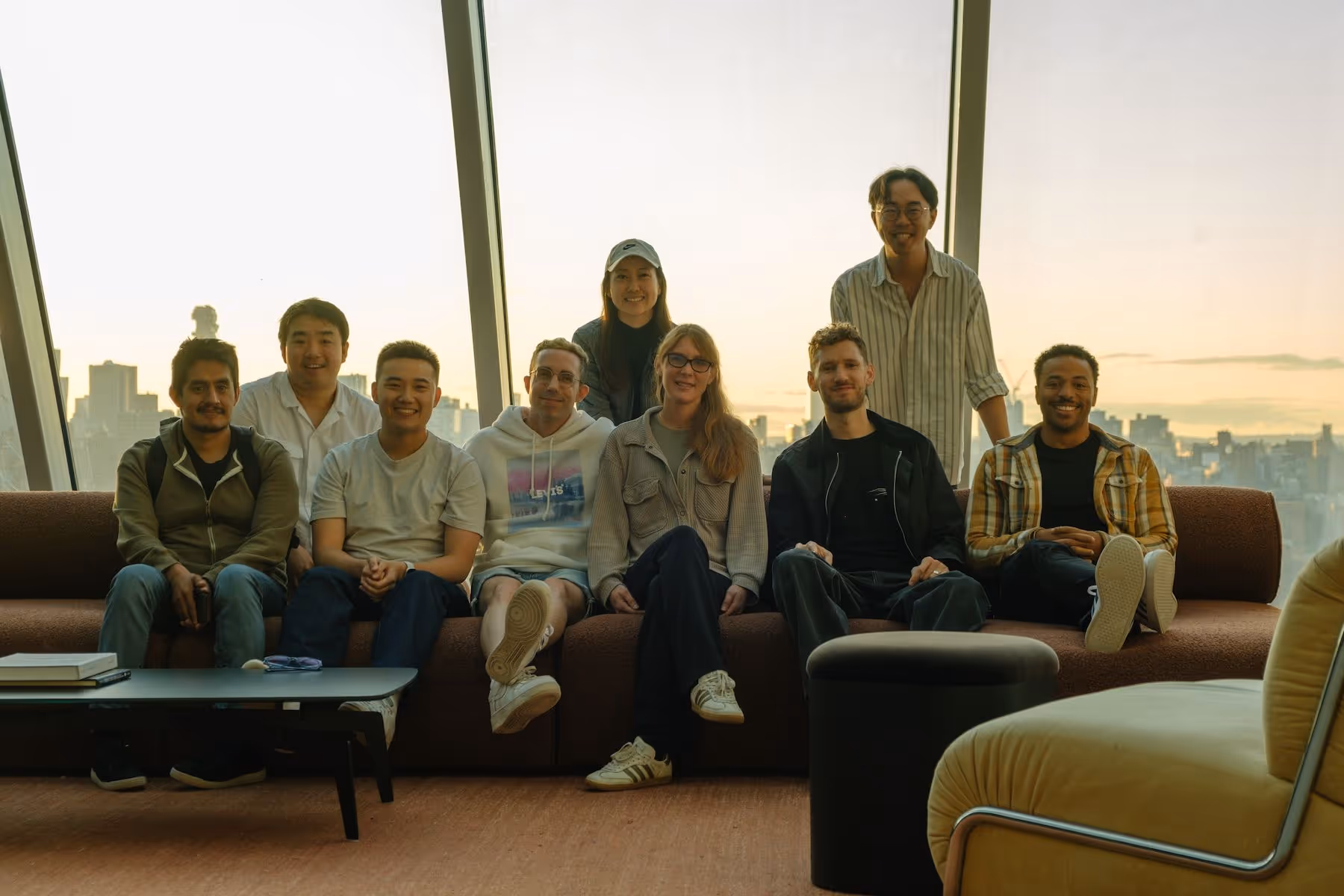 Group of nine young adults smiling and sitting on a couch in a room with large windows and city view