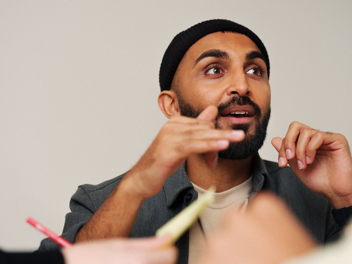 Homme barbu portant un bonnet noir et une chemise grise, s'exprimant avec des gestes pendant une discussion.