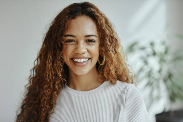 Une femme souriante aux cheveux longs et bouclés porte un pull blanc avec un arrière-plan flou d'intérieur.