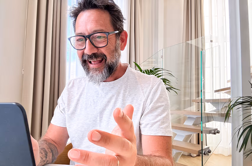 Homme souriant avec lunettes et barbe discutant devant un ordinateur portable dans une pièce lumineuse avec plantes et fenêtres hautes.