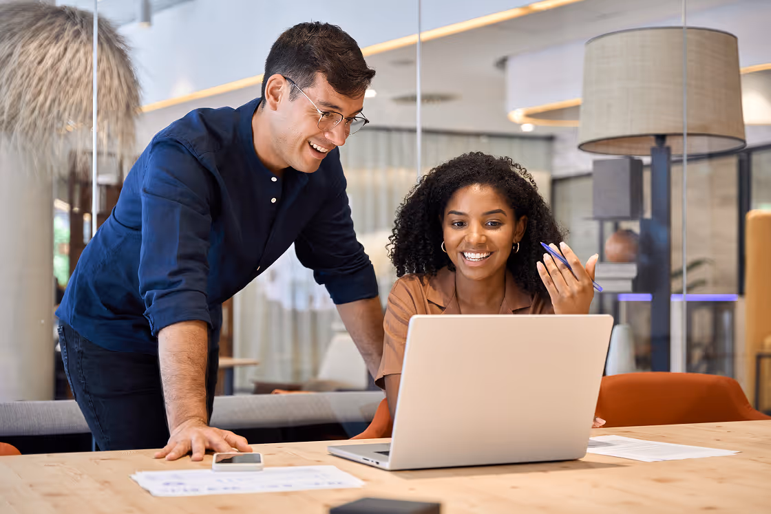 Deux collègues souriants travaillant ensemble sur un ordinateur portable dans un bureau moderne.